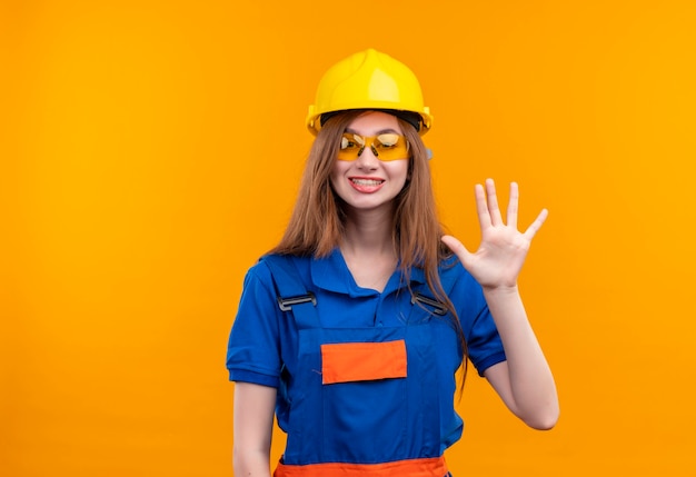 Młoda kobieta pracownik budowniczy w mundurze budowy i hełmie ochronnym uśmiechnięty pokazując i wskazując palcami numer pięć stojących na pomarańczowej ścianie