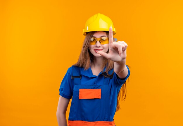 Młoda kobieta pracownik budowniczy w mundurze budowy i kask ochronny uśmiechnięty pokazując palec wskazujący z przodu