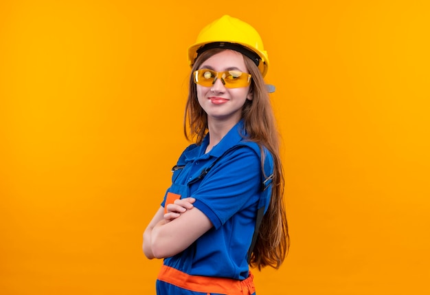 Młoda kobieta pracownik konstruktora w mundurze budowy i hełm ochronny stojący z rękami skrzyżowanymi patrząc pewnie na pomarańczową ścianę