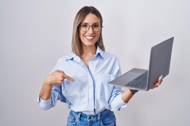 Młoda kobieta pracująca przy użyciu laptopa komputerowego, wyglądająca pewnie z uśmiechem na twarzy, wskazująca na siebie palcami dumnymi i szczęśliwymi