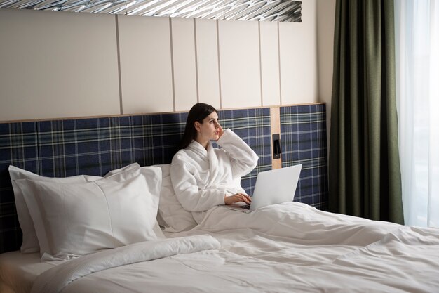 Młoda kobieta pracuje na swoim laptopie, siedząc na łóżku w pokoju hotelowym