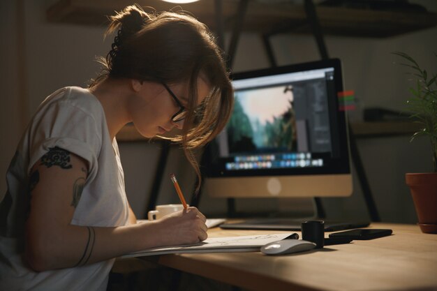 Młoda kobieta projektant siedzi w pomieszczeniu w nocy, rysując szkice