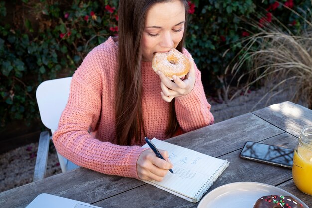 Młoda kobieta robi notatki na zewnątrz podczas jedzenia pączka