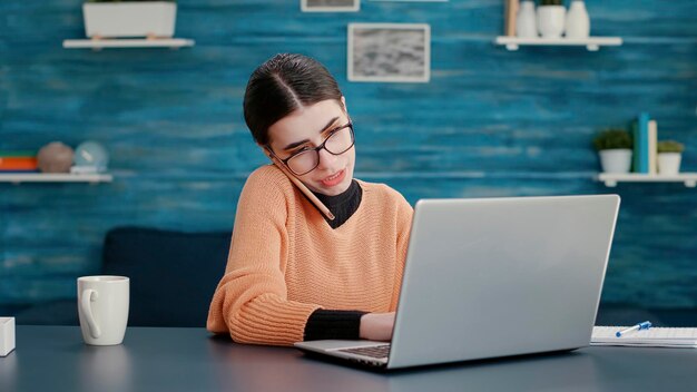 Młoda kobieta rozmawia przez telefon i korzysta z laptopa na biurku w domu, uczęszczając na lekcję online do zdalnej edukacji. Nowoczesna komunikacja na smartfonie, praca w szkole na komputerze.