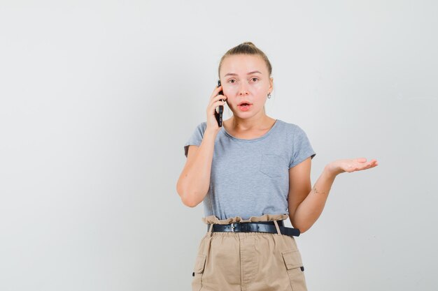 Młoda kobieta rozmawia przez telefon komórkowy w t-shirt, spodnie i patrząc zaskoczony. przedni widok.