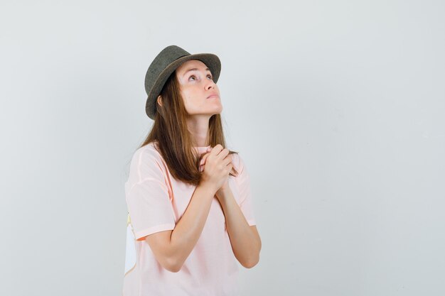 Młoda kobieta, ściskając ręce w geście modlitwy w różowej koszulce, kapeluszu i patrząc z nadzieją. przedni widok.
