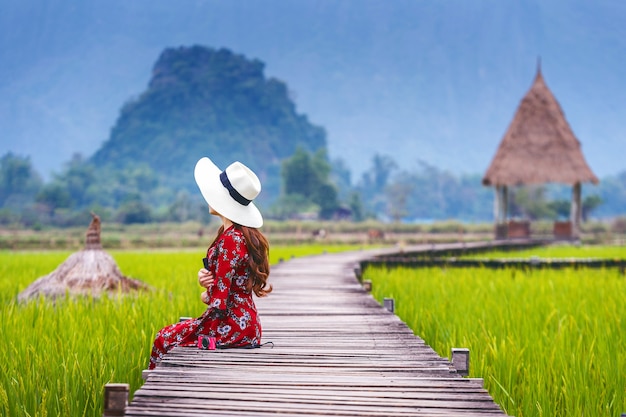 Bezpłatne zdjęcie młoda kobieta siedzi na drewnianej ścieżce z zielonym ryżowym polem w vang vieng, laos.