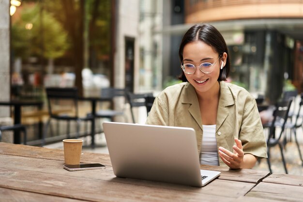 Młoda kobieta siedzi na spotkaniu online w kawiarni na świeżym powietrzu, rozmawiając z kamerą laptopa, wyjaśniając coś