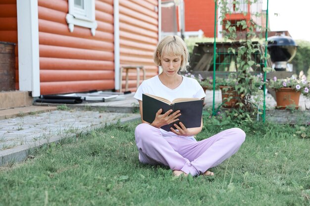 Młoda kobieta siedzi na trawie na podwórku w słoneczny dzień i czyta prawdziwą książkę. trawa i mniszek tło, ładny wygląd vintage.