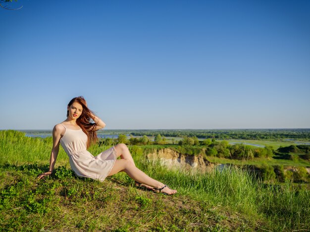 Młoda kobieta siedzi przy klifie na świeżym powietrzu na charakter. Atrakcyjna dziewczyna w białej sukni pozowanie na świeżym powietrzu. Modelka stwarzających w polu w słoneczny letni dzień.
