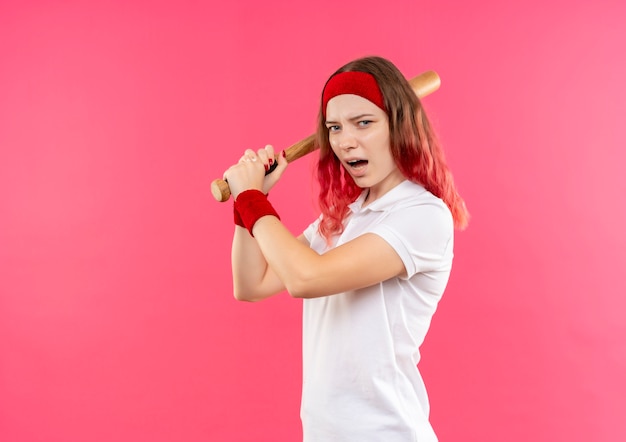Młoda kobieta sportowa w opasce kołysząc kijem, grając w baseball stojąc na różowej ścianie