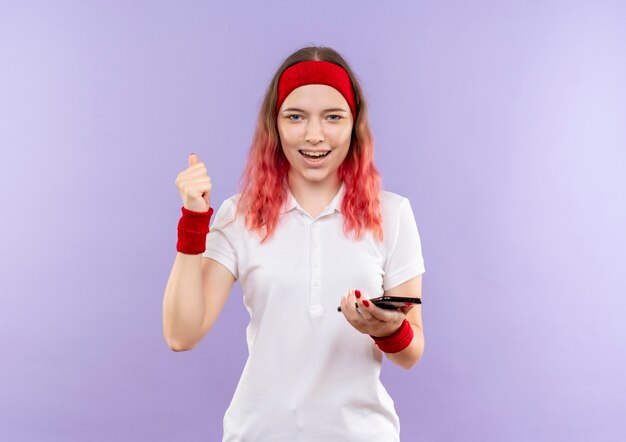 Młoda kobieta sportowy trzymając smartfon zaciskając pięść uśmiechnięty radośnie stojąc nad fioletową ścianą