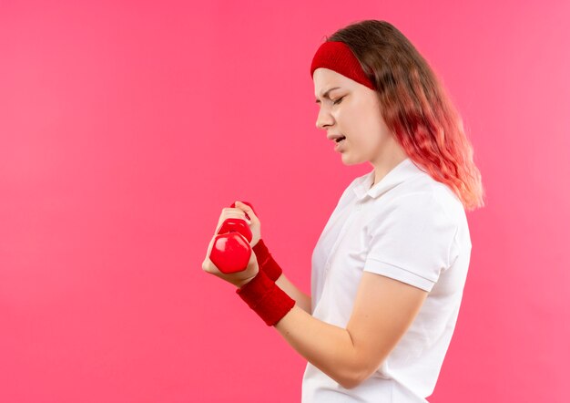 Młoda kobieta sportowy w opasce robi ćwiczenia z hantlami patrząc napięty stojącego na różowej ścianie