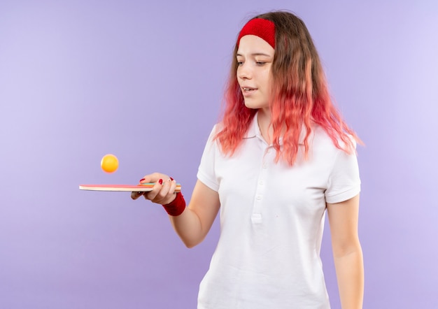 Młoda kobieta sportowy w opasce trzyma rakietę do tenisa stołowego i piłki, grając z uśmiechem na twarzy stojącej nad fioletową ścianą