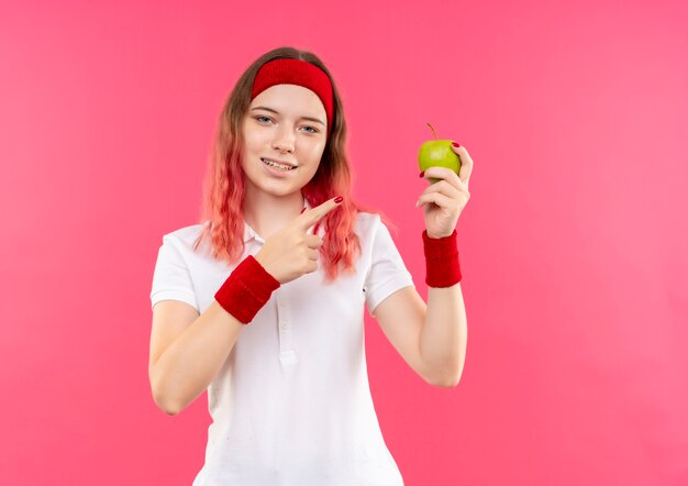 Młoda kobieta sportowy w opasce trzyma zielone jabłko, wskazując palcem na to, uśmiechając się pewnie stojąc na różowej ścianie