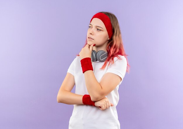 Młoda kobieta sportowy ze słuchawkami, patrząc na bok z ręką na brodzie myśli stojącej nad fioletową ścianą