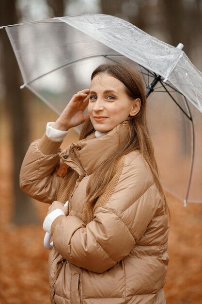 Młoda kobieta stojąca w jesiennym lesie Brunetka kobieta trzymająca przezroczysty parasol Dziewczyna ubrana w modną brązową kurtkę