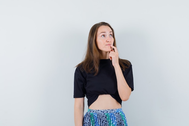 Młoda kobieta stojąca w myśleniu poza i patrząc w czarny t-shirt i niebieską spódnicę i patrząc zamyślony