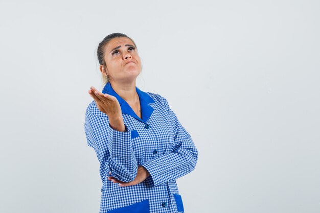 Młoda kobieta stojąca w myśleniu poza w niebieskiej kraciastej koszuli piżamy i patrząc zmęczony, widok z przodu.
