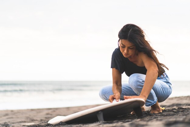 Młoda kobieta surfer przygotowuje deskę surfingową na oceanie, woskowanie Kobieta z deską surfingową na oceanie aktywny styl życia sporty wodne