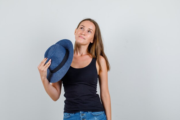 Młoda kobieta trzyma kapelusz podczas pozowanie w podkoszulku, widok z przodu szorty.