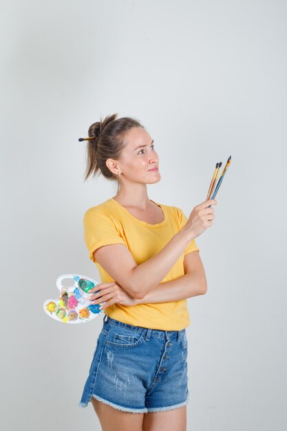 Młoda kobieta trzyma paletę sztuki ze szczotkami w żółtej koszulce, dżinsach i patrząc szczęśliwy