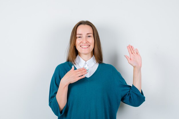 Młoda kobieta trzyma rękę na klatce piersiowej, pokazując dłoń w swetrze na białej koszuli i patrząc wesoło. przedni widok.
