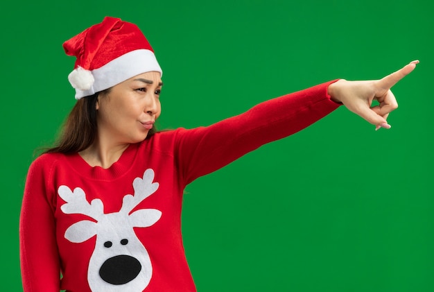 Młoda kobieta ubrana w świąteczny santa hat i czerwony sweter patrząc na bok z poważną twarzą wskazującą palcem wskazującym stojącym na zielonym tle