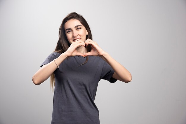 Młoda kobieta ubrana w szary t-shirt na szarym tle robi kształt symbolu serca rękami.