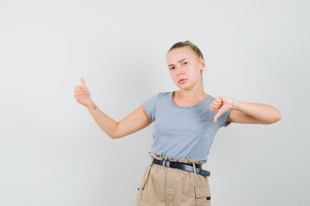 Bezpłatne zdjęcie młoda kobieta udająca, że pokazuje coś z kciukiem w dół w koszulce, spodniach i wygląda na rozczarowaną. przedni widok.
