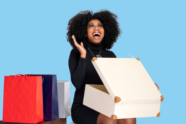 Młoda kobieta uśmiech się szczęśliwie, otwierając pudełko z prezentami, siadając i obok kilku toreb na zakupy
