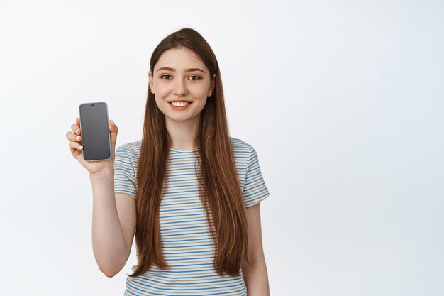 Młoda kobieta uśmiechający się zadowolony, pokazując smartfon pusty ekran, interfejs aplikacji. Koncepcja sklepu mobilnego, bankowości internetowej lub zakupów, białe tło.
