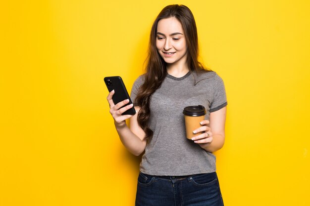 Młoda kobieta używa telefonu trzymając filiżankę kawy na białym tle na żółtej ścianie