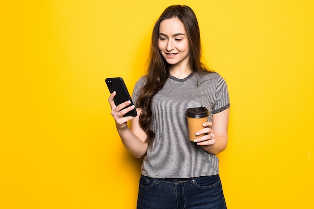 Młoda kobieta używa telefonu trzymając filiżankę kawy na białym tle na żółtej ścianie