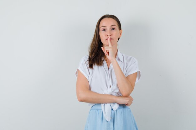 Młoda kobieta w białej bluzce i jasnoniebieskiej spódnicy pokazuje gest ciszy i wygląda poważnie