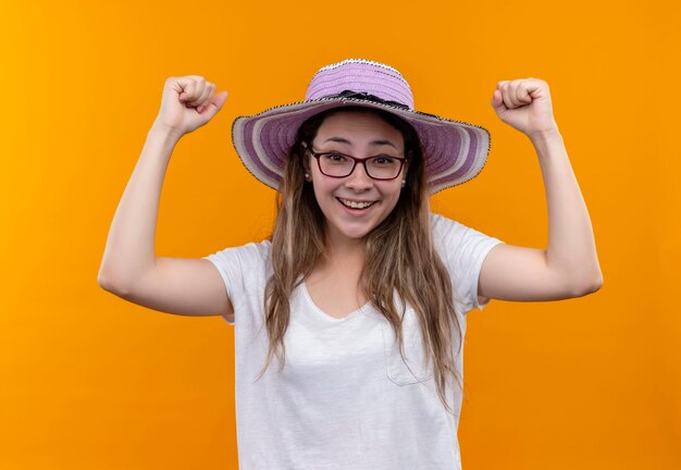 Młoda kobieta w białej koszulce w letnim kapeluszu, podnosząc pięści, szczęśliwa i podekscytowana, ciesząc się swoim sukcesem stojąc nad pomarańczową ścianą