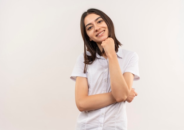 Młoda kobieta w białej koszuli patrząc do przodu uśmiechnięta pewna siebie stojąca na białej ścianie