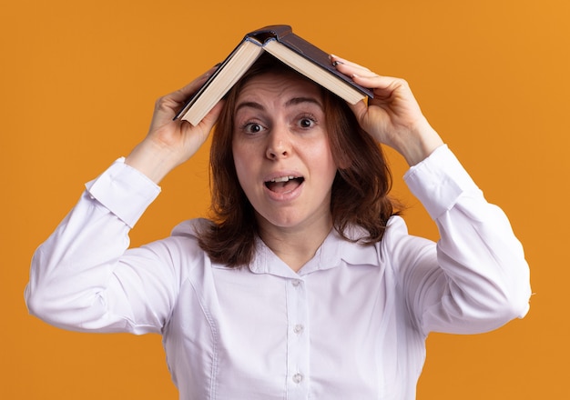 Młoda kobieta w białej koszuli, trzymając otwartą książkę nad głową, uśmiechając się zdezorientowany stojąc nad pomarańczową ścianą