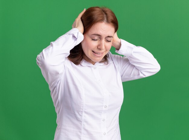 Młoda kobieta w białej koszuli wygląda źle, dotykając jej głowy cierpiącej na ból głowy stojącej nad zieloną ścianą