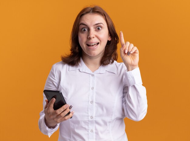 Młoda kobieta w białej koszuli ze smartfonem patrząc z przodu z uśmiechem na twarzy pokazując palec wskazujący o nowym pomyśle stojącym nad pomarańczową ścianą