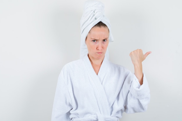 Młoda kobieta w białym szlafroku, ręcznik wskazując kciukiem i patrząc wątpliwie, widok z przodu.
