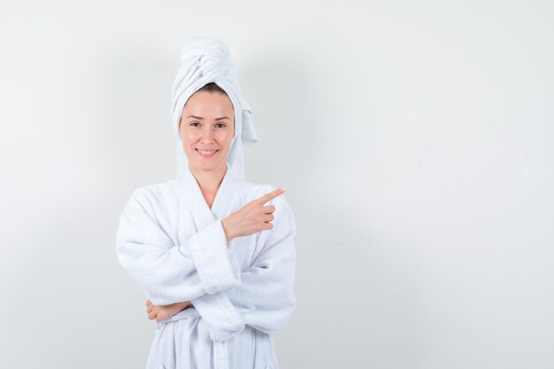 Młoda kobieta w białym szlafroku, ręcznik, wskazując w prawym górnym rogu i patrząc wesoły, przedni widok.