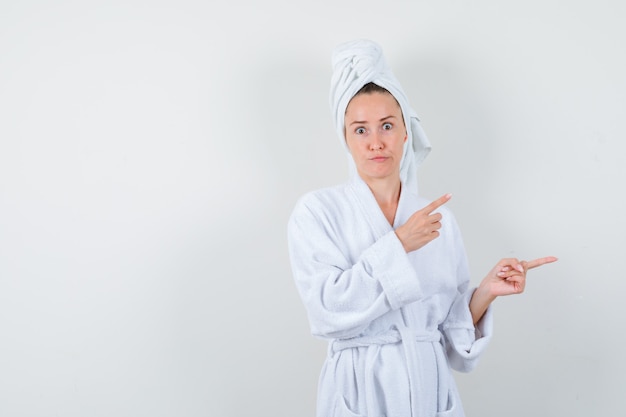 Młoda kobieta w białym szlafroku, ręcznik wskazujący w prawo i patrząc zdziwiony, widok z przodu.