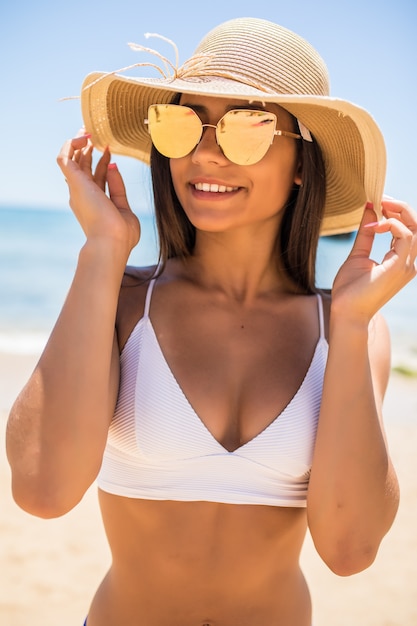 Młoda kobieta w bikini na sobie biały słomkowy kapelusz, ciesząc się wakacjami na plaży. Portret pięknej kobiety Łacińskiej relaks na plaży z okularami przeciwsłonecznymi.