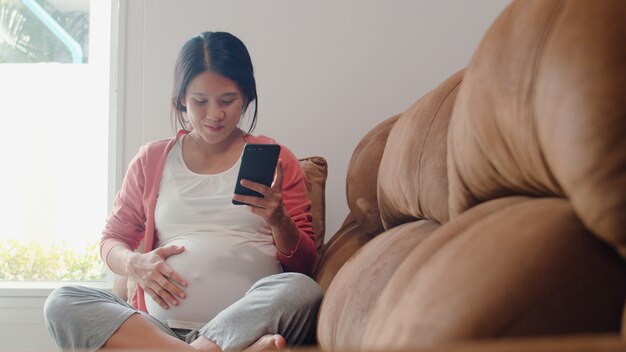 Młoda kobieta w ciąży Azji przy użyciu telefonu komórkowego wyszukiwania informacji o ciąży. Mama czuje się szczęśliwy, uśmiechnięty pozytywnie i spokojnie, podczas gdy dbać o swoje dziecko leżące na kanapie w salonie w domu.