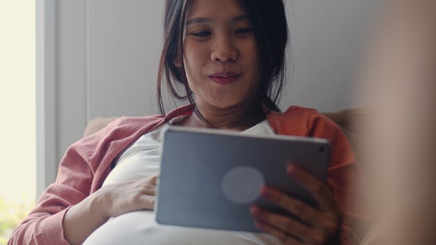 Młoda kobieta w ciąży Azji za pomocą tabletu wyszukiwania informacji o ciąży. Mama czuje się szczęśliwy, uśmiechnięty pozytywnie i spokojnie, podczas gdy dbać o swoje dziecko leżące na kanapie w salonie w domu.