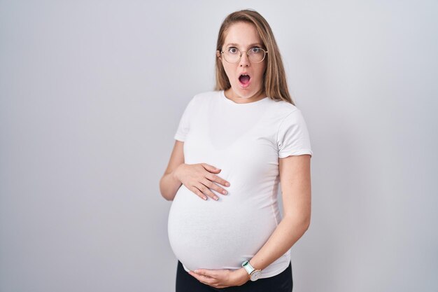 Młoda kobieta w ciąży spodziewająca się dziecka, dotykająca ciężarnego brzucha przestraszona i zszokowana zaskoczeniem i zdumionym wyrazem twarzy, strachem i podekscytowaną twarzą.