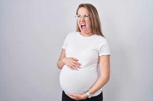 Młoda kobieta w ciąży spodziewająca się dziecka, dotykająca ciężarnego brzucha wściekła i szalona, krzycząca sfrustrowana i wściekła, krzycząca ze złości. wściekłość i agresywna koncepcja.