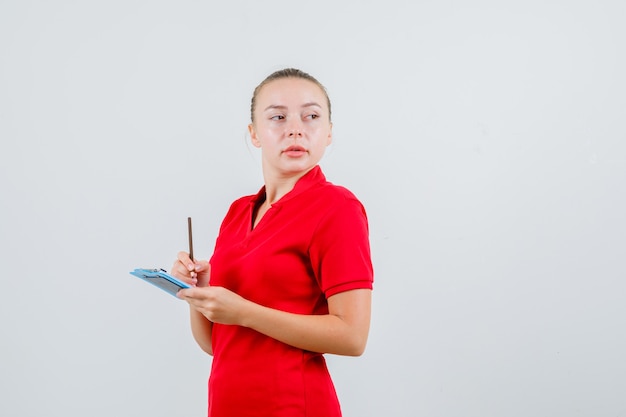 Bezpłatne zdjęcie młoda kobieta w czerwonej koszulce trzymając schowek i ołówek i patrząc zamyślony