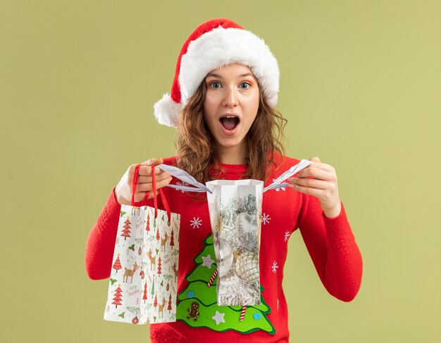 Młoda kobieta w czerwonym świątecznym swetrze i santa hat trzyma papierowe torby z prezentami świątecznymi otwierając torby będąc szczęśliwa i zaintrygowana stojąc nad zieloną ścianą